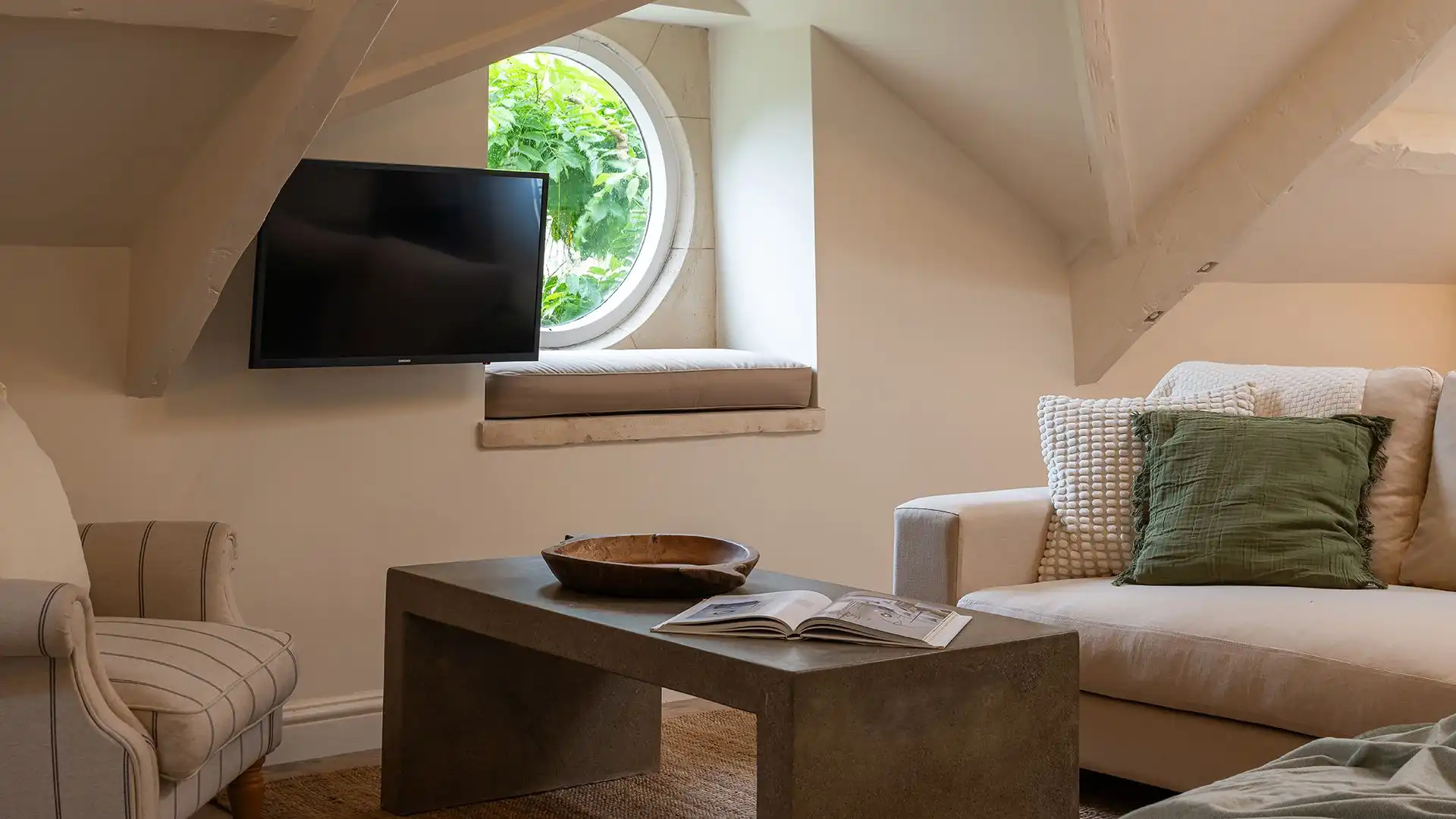 There is a coffee table, wall-mounted smart TV and a cushioned window seat beside a circular statement window.